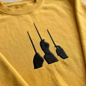 May include: A mustard yellow crewneck sweatshirt featuring a black embroidered design on the chest. The embroidery depicts three silhouettes of hands holding thin wands or sticks, positioned vertically in a row against the solid yellow fabric.