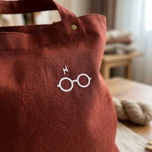 Peut inclure: Un sac fourre-tout en toile couleur rouille orné d'une broderie blanche représentant des lunettes rondes surmontées d'un éclair. Le sac est doté d'une anse robuste et d'un petit rivet métallique situé près du bord supérieur du tissu.