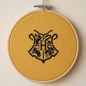 May include: A circular wooden embroidery hoop containing mustard yellow fabric with a black embroidered crest featuring a central letter H surrounded by four distinct animal figures, suspended by a thin piece of twine from the top metal screw.