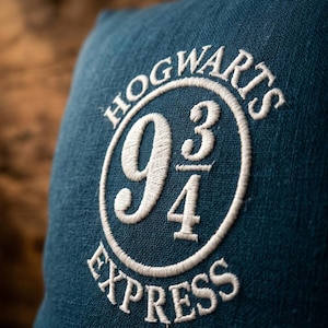 May include: A dark teal square cushion featuring white embroidered text that reads HOGWARTS EXPRESS in a circular pattern around the number 9 3/4. The fabric has a visible woven texture and the embroidery is stitched in a clean, white thread.