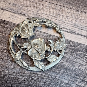 May include: A silver-colored, circular decorative piece featuring a detailed floral design. The piece showcases roses and leaves arranged in a circular pattern. The metal has a slightly aged appearance, adding to its vintage charm. The design is intricate and well-defined.