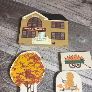 May include: Wooden fall-themed decorative cutouts. Includes a house with "BASKETMAKER" text, a tree with a swing, a wagon filled with pumpkins, and a scene with turkeys and pumpkins. The house has a black cat cutout.