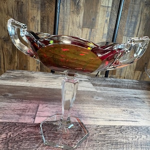 May include: A clear glass pedestal bowl with a ruby red and gold interior. The bowl has two handles and sits on a clear, faceted stem and base. The bowl is approximately 12 inches wide.