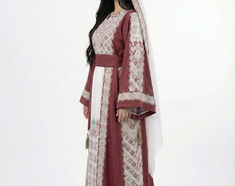 May include: A long, maroon dress with white and embroidered details. The dress features long sleeves, a matching belt, and a white head covering. The embroidery includes geometric and floral patterns. Tassels hang from the dress.