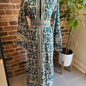 May include: A teal-colored, long-sleeved dress with intricate cream-colored floral embroidery. The dress features a matching belt and a high neckline. The embroidery is detailed and covers the entire dress, creating a textured appearance.