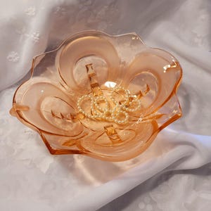 Peut inclure: Un bol en verre translucide de couleur pêche avec un bord festonné en forme de fleur et un motif moulé de segments incurvés rayonnant depuis le centre. Un délicat collier de petites perles artificielles crème repose dans le fond.