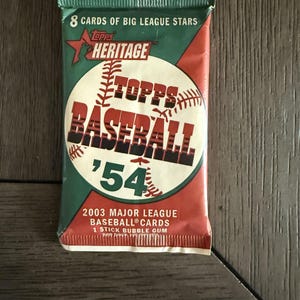 May include: A sealed pack of 2003 Topps Heritage baseball cards featuring a green and red wrapper with a baseball graphic. The text reads 8 CARDS OF BIG LEAGUE STARS, TOPPS HERITAGE, TOPPS BASEBALL '54, 2003 MAJOR LEAGUE BASEBALL CARDS, and 1 STICK BUBBLE GUM.