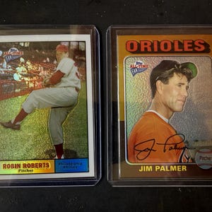May include: Two baseball cards in protective cases. One features Robin Roberts, pitcher for the Philadelphia Phillies. The other card displays Jim Palmer of the Orioles. Both cards are from the All-Time Fan Favorites series.