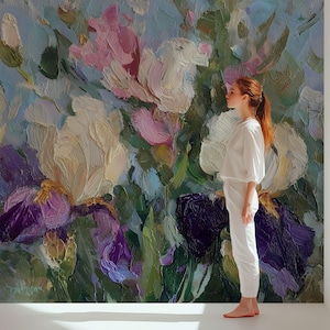 May include: Oil painting of large irises in shades of purple, cream, and pink, with green foliage against a light blue background. A person in white clothing stands in front of the painting.