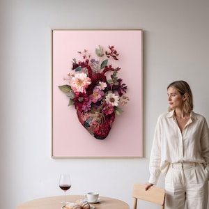 May include: A framed artwork featuring a stylized anatomical heart adorned with a vibrant floral arrangement. The heart is a deep burgundy color, and the flowers include shades of pink, red, and white. The background is a soft pink, and the frame is a light wood color.