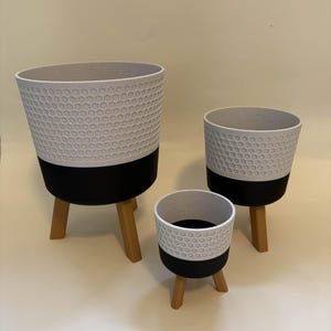 Puede incluir: Conjunto de tres macetas modernas. Las macetas tienen una sección superior blanca con textura de panal, una sección inferior negra y están soportadas por tres patas de madera. Los tamaños varían, ofreciendo versatilidad para diferentes plantas.