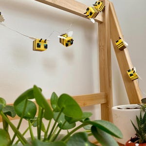 May include: A string of decorative lights featuring yellow and black bee-shaped ornaments. The lights are strung on a thin wire and are displayed on a wooden frame. The background includes green plants in white and red pots.