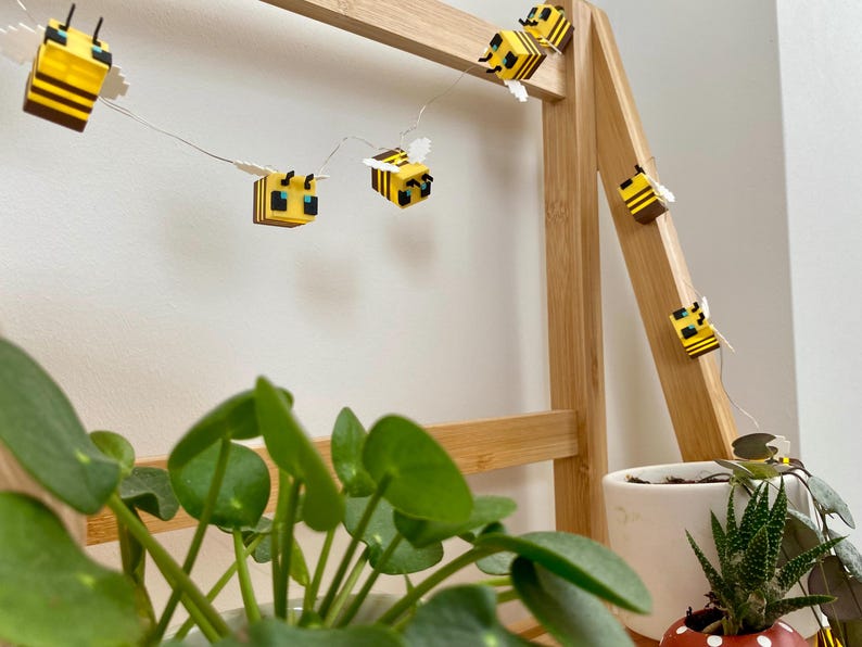 May include: A string of decorative lights featuring yellow and black bee-shaped ornaments. The lights are strung on a thin wire and are displayed on a wooden frame. The background includes green plants in white and red pots.