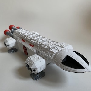 Può includere: Un'astronave giocattolo bianca con un cono nasale nero e ugelli motore rossi. Il modello presenta una struttura a traliccio dettagliata sulla parte superiore e carrello di atterraggio grigio. Il giocattolo è una replica di un'astronave.