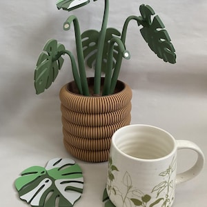以下が含まれることがあります： 緑の葉と茶色の鉢植えの装飾的な木製の植物が、緑の葉のデザインが施された白いマグカップの隣に置かれています。緑、白、グレーのアクセントが付いた葉の形をしたコースターも存在します。