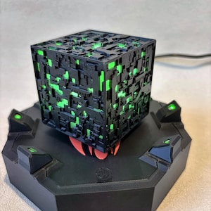 May include: A black cube with a complex, mechanical surface texture featuring glowing green light emanating from within. The cube rests on a black hexagonal base equipped with four small, upward-facing green lights at each corner of the platform.