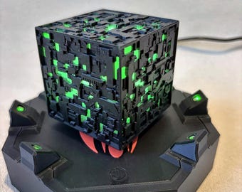 Può includere: Un cubo nero con una complessa trama superficiale meccanica da cui emana una luce verde brillante. Il cubo poggia su una base esagonale nera dotata di quattro piccole luci verdi rivolte verso l'alto posizionate su ogni angolo della piattaforma.