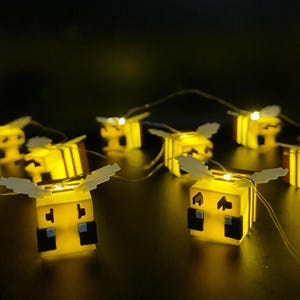 May include: A string of yellow bee-shaped LED lights with white wings. The lights are arranged on a thin wire, creating a whimsical and decorative lighting display. The bees have black and blue accents.