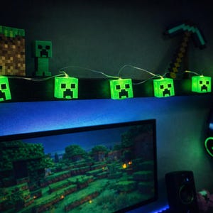 Puede incluir: Una habitación temática de Minecraft con luces de cadena verdes brillantes con caras de Creeper. También se ven un pico pixelado y un bloque de Minecraft. Un monitor de computadora muestra un paisaje de Minecraft.