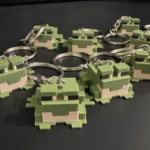 Puede incluir: Un conjunto de llaveros de rana pixelados verdes y beige. Cada llavero presenta un pequeño diseño de rana en bloques con un llavero plateado. Las ranas están en varias posiciones, sobre un fondo oscuro.
