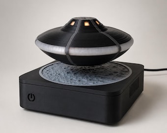 Può includere: Una lampada a forma di UFO in levitazione con esterno nero, una fascia bianca illuminata e piccole finestre luminose. L'oggetto fluttua sopra una base quadrata nera con una superficie testurizzata e craterizzata simile alla luna.