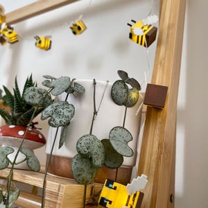 May include: A wooden ladder shelf displays potted plants and a string of pixelated bee lights. The plants include a succulent in a mushroom-shaped pot and a trailing plant with heart-shaped leaves. The bee lights are yellow and black with white wings.