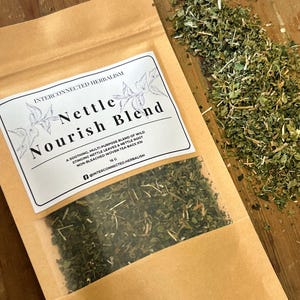 Könnte beinhalten: Ein brauner Papiertüte mit einem transparenten Fenster, das eine grüne Kräutermischung enthält. Das Etikett lautet "Nettle Nourish Blend" mit Details zu den Inhaltsstoffen und der Marke "Interconnected Herbalism". Lose Kräuter sind neben der Tüte verstreut.