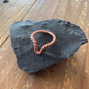 Pode incluir: Um anel de cor cobre com um design de corda torcida. O anel está sobre uma pedra cinza escura de formato irregular, que repousa sobre uma superfície de madeira. O design do anel apresenta um padrão trançado contínuo.