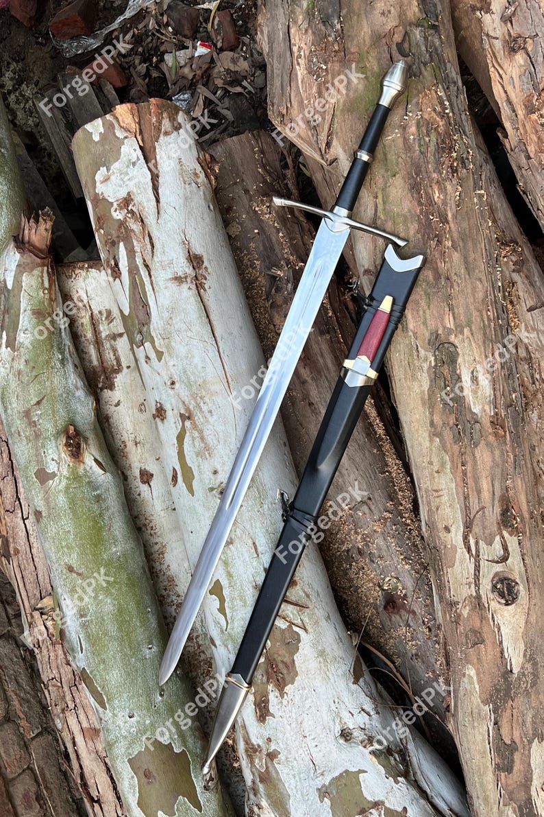May include: A silver sword with a black handle and scabbard. The sword is lying on a pile of wooden logs.