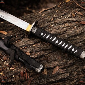 May include: A Japanese-style sword with a silver blade and a black handle wrapped with white diamond-shaped accents. The sword's handle and sheath are black, with decorative silver accents. The sword is resting on a textured, brown wooden surface.