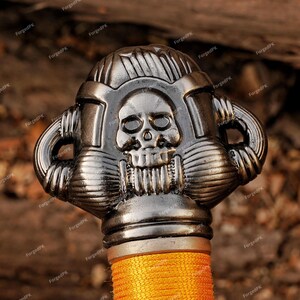 May include: Close-up of a decorative sword handle with a skull design. The handle is silver-colored with intricate details and a textured appearance. The grip is wrapped in orange cord.