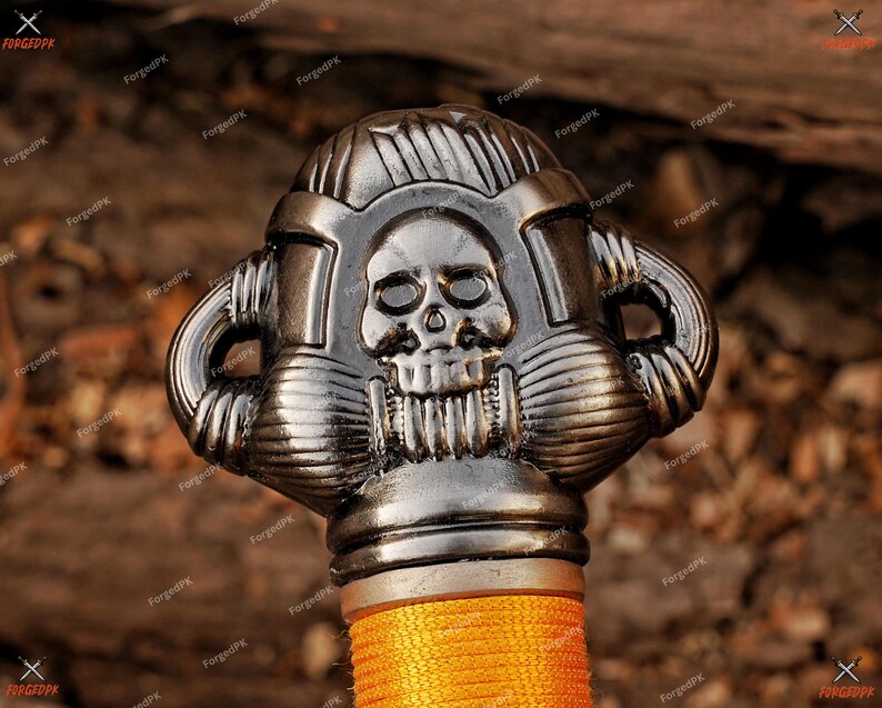 May include: Close-up of a decorative sword handle with a skull design. The handle is silver-colored with intricate details and a textured appearance. The grip is wrapped in orange cord.