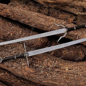 May include: Two steel swords with silver blades and black handles rest on a bed of wood. One sword has a unique, forked crossguard, while the other features a circular emblem on the hilt. The swords are likely for display or costume purposes.