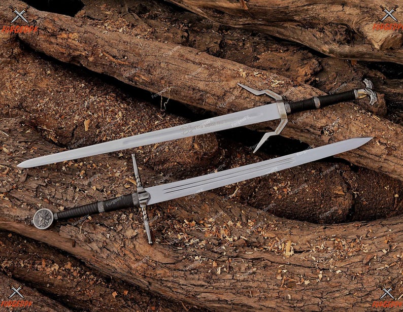 May include: Two steel swords with silver blades and black handles rest on a bed of wood. One sword has a unique, forked crossguard, while the other features a circular emblem on the hilt. The swords are likely for display or costume purposes.