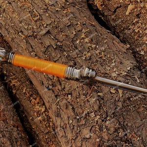 May include: A decorative dagger with a silver handle and a bright orange wrapped grip. The blade is silver and the dagger rests on a bed of brown wood.