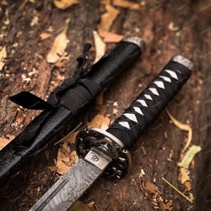 May include: Close-up of a katana sword with a black handle and scabbard. The blade has a unique pattern, and the handle is wrapped with white and black material. The sword rests on a textured, brown surface.