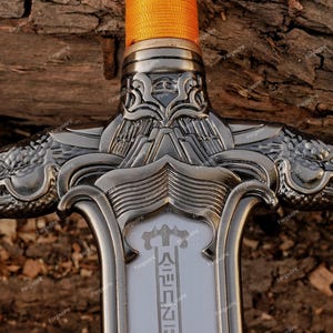 May include: Close-up of a decorative sword with a silver-colored blade and handle. The handle features two dragon head designs on either side. The sword's grip is wrapped in orange fabric. The blade has text in a foreign language.