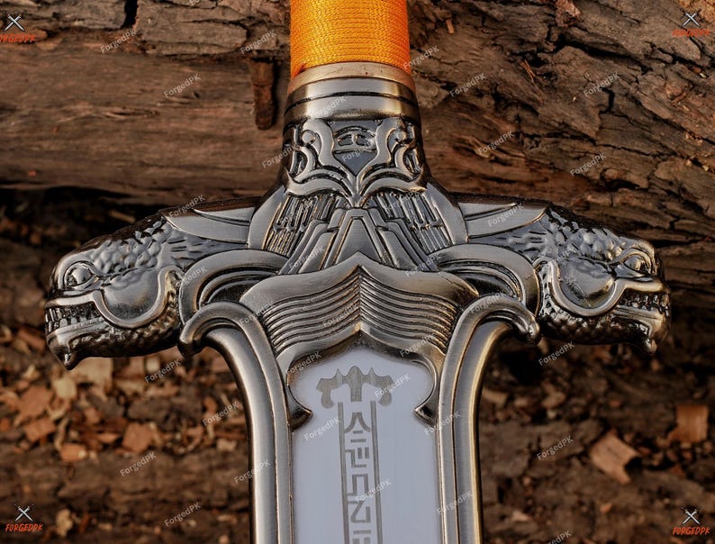 May include: Close-up of a decorative sword with a silver-colored blade and handle. The handle features two dragon head designs on either side. The sword's grip is wrapped in orange fabric. The blade has text in a foreign language.