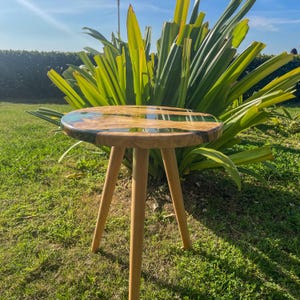 May include: A small, round wooden table with a green and brown epoxy resin top. The table has three legs and is sitting on a grassy lawn.