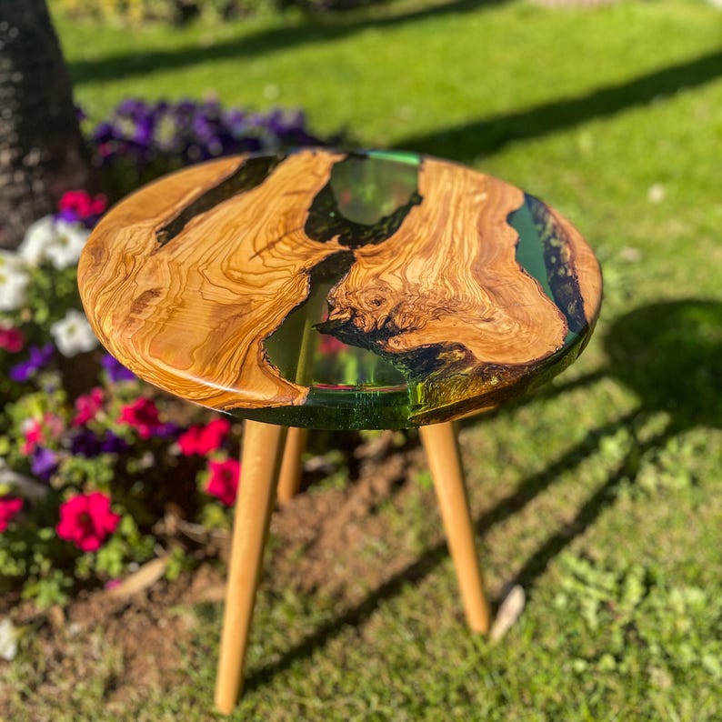 May include: A round wooden table with a green and black epoxy resin inlay. The table has three wooden legs and is sitting on a grassy surface.