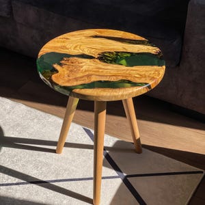 May include: A round wooden side table with three legs. The tabletop is made of wood with a green epoxy resin inlay.