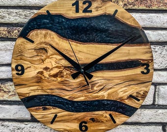 May include: A round wooden wall clock with a natural wood grain pattern and dark blue resin accents. The clock features black hands and black numerals at the 12, 3, 6, and 9 o'clock positions. The clock is mounted on a brick wall.