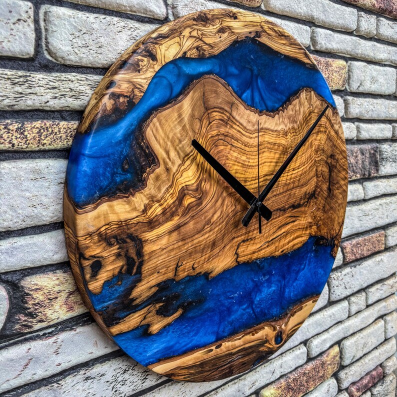 Puede incluir: Un reloj de pared redondo de madera con un dise&ntilde;o &uacute;nico. La esfera del reloj presenta vetas de madera natural con un r&iacute;o de resina azul vibrante. Las manecillas negras indican la hora. El reloj est&aacute; montado en una pared de ladrillo.