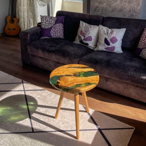 May include: A round wooden side table with a green epoxy resin top. The table has three legs and is sitting on a white rug with black lines.
