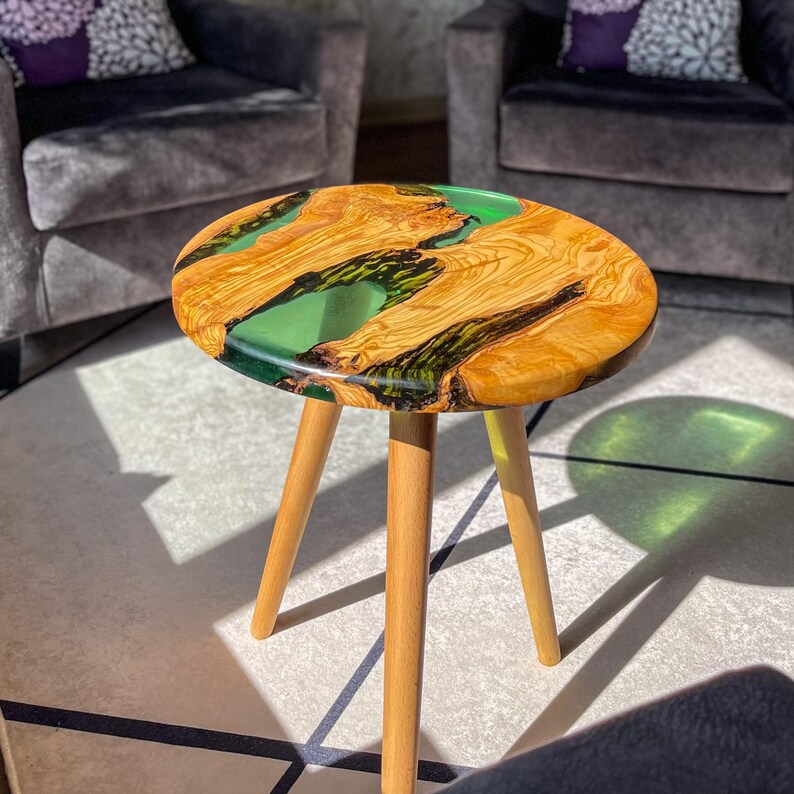 May include: A round wooden side table with three legs. The tabletop is made of wood with a green epoxy resin inlay.