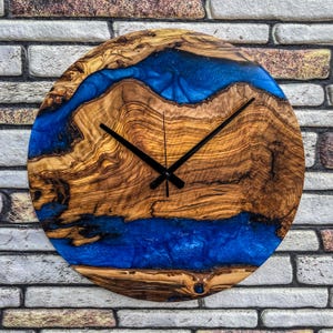 Puede incluir: Reloj de pared redondo de madera con un dise&ntilde;o &uacute;nico. La esfera del reloj presenta vetas de madera natural con un dise&ntilde;o de r&iacute;o de resina azul vibrante. Las manecillas negras indican la hora. El reloj est&aacute; montado en una pared de ladrillo.