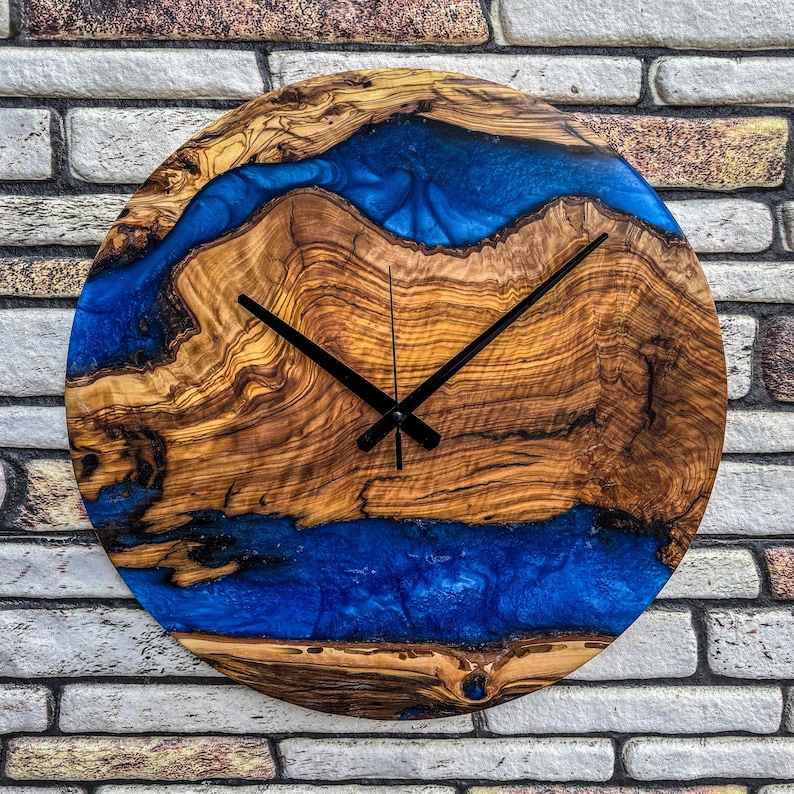 Puede incluir: Reloj de pared redondo de madera con un dise&ntilde;o &uacute;nico. La esfera del reloj presenta vetas de madera natural con un dise&ntilde;o de r&iacute;o de resina azul vibrante. Las manecillas negras indican la hora. El reloj est&aacute; montado en una pared de ladrillo.