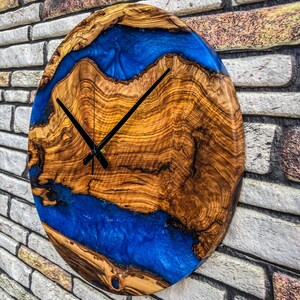 Puede incluir: Reloj de pared ovalado de madera con un patr&oacute;n de vetas de madera natural y un dise&ntilde;o de r&iacute;o de resina azul vibrante. El reloj tiene manecillas negras y est&aacute; montado en una pared de ladrillo. El di&aacute;metro del reloj es de aproximadamente 30 cm.