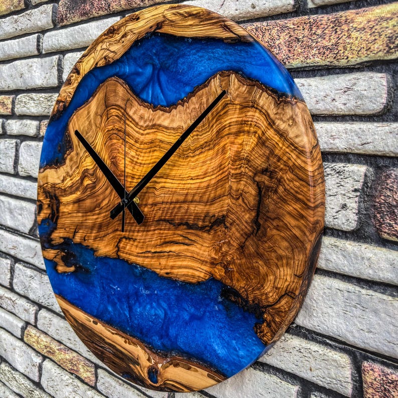 Puede incluir: Reloj de pared ovalado de madera con un patr&oacute;n de vetas de madera natural y un dise&ntilde;o de r&iacute;o de resina azul vibrante. El reloj tiene manecillas negras y est&aacute; montado en una pared de ladrillo. El di&aacute;metro del reloj es de aproximadamente 30 cm.