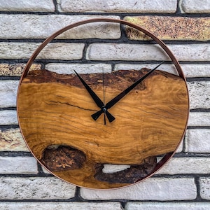 Pode incluir: Um relógio de parede redondo de madeira com uma moldura de metal de cor cobre. O mostrador do relógio é feito de uma fatia de madeira natural com um padrão de grão único. Os ponteiros do relógio são pretos.